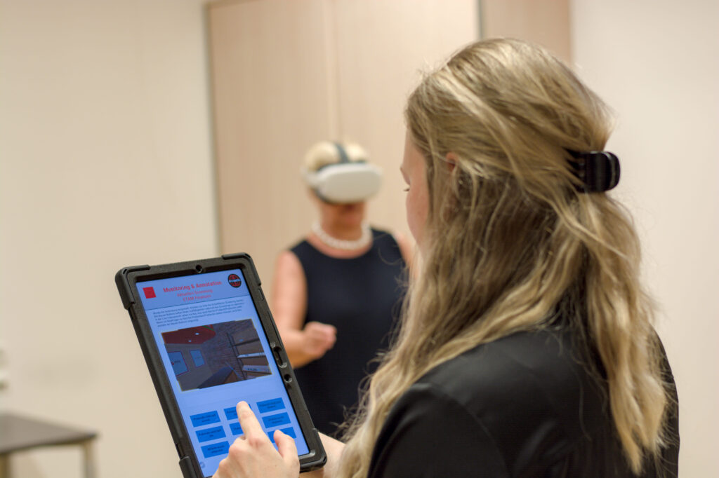 Eine Frau bedient ein Tablet, auf dem die Live-Übertragung der VR-Brille und Buttons zu sehen sind. Im Hintergrund steht eine weitere Frau, die eine VR-Brille trägt und eine Aufgabe in der virtuellen Realität erfüllt.